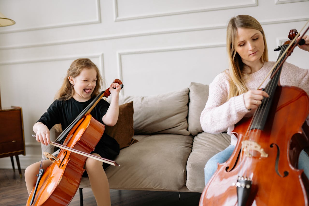 A woman and girl joyfully play cellos together in a cozy living room setting.